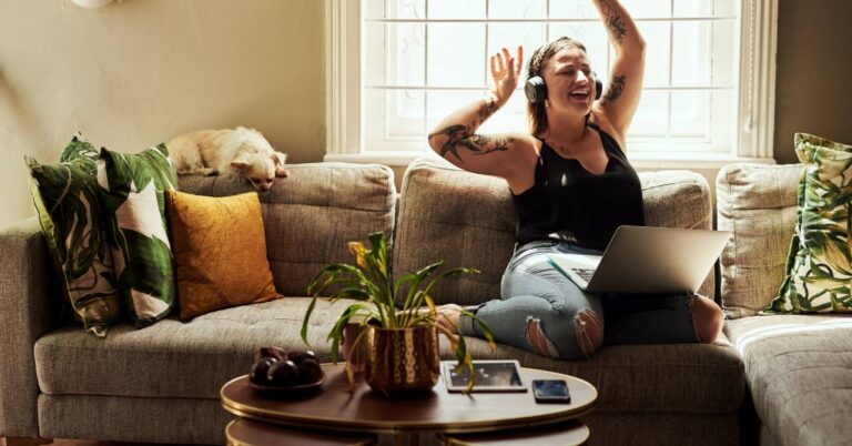 Woman sitting on a sofa with a laptop and headphones listening music happy dancing — daily habits for happiness